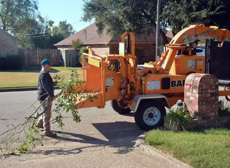 Groundman feeding a leafy limb into an orange towable brush chipper parked in a suburban driveway